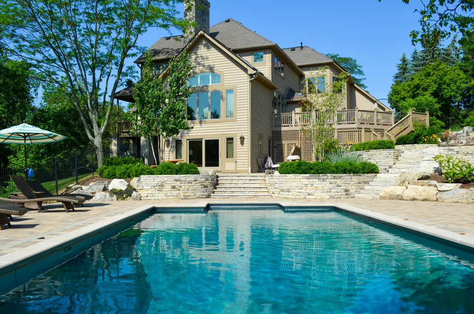 Sided house overlooking a rectangular pool with paver pool deck, lounge chairs, umbrella, limestone walls, and upper deck.